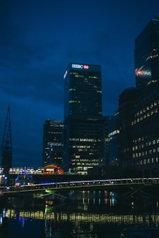 A night-time cityscape featuring a well-lit skyscraper with an HSBC logo prominently displayed at the top. The scene includes several tall buildings with lights in their windows reflecting off a body of water below. A bridge, also illuminated, spans across the water in the foreground.
