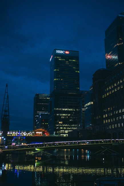A night-time cityscape featuring a well-lit skyscraper with an HSBC logo prominently displayed at the top. The scene includes several tall buildings with lights in their windows reflecting off a body of water below. A bridge, also illuminated, spans across the water in the foreground.