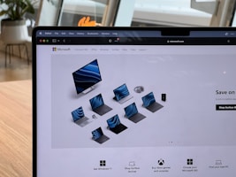 A laptop screen displays the Microsoft website featuring a range of Surface products, including a monitor, laptops, tablets, and headphones. The website is being viewed through the Safari browser. The background shows a modern indoor setting with wooden furniture and large windows.