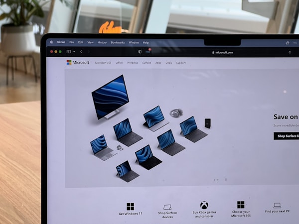A laptop screen displays the Microsoft website featuring a range of Surface products, including a monitor, laptops, tablets, and headphones. The website is being viewed through the Safari browser. The background shows a modern indoor setting with wooden furniture and large windows.