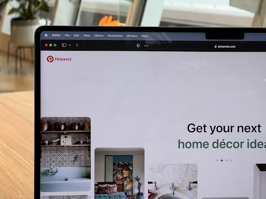 A laptop screen displaying the Pinterest website. The web page shows various home decor ideas with images of decorative items and interiors. The laptop is placed on a wooden surface in a room with plants and modern furniture in the background.