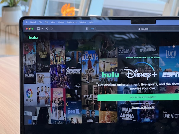 A laptop screen displays a web page featuring a mix of movie and TV show covers, highlighting popular titles and a Hulu and Disney+ bundle promotion. There are visible icons and images of films and shows alongside promotional text offering entertainment and live sports.