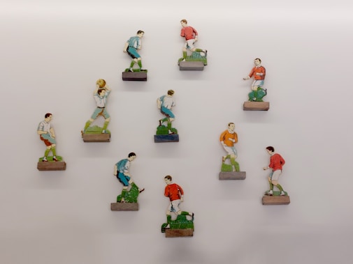 A collection of painted wooden figurines depicting soccer players mounted on small blocks, arranged in a grid-like pattern on a light-colored wall. Each figurine is in a dynamic pose, suggesting movement typical of a soccer game. The players are wearing uniforms in red, blue, and orange, and their hairstyles and expressions are distinct.