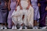 Five women are standing in a line, wearing elegant dresses in shades of purple and lavender with silver sneakers. The central figure is wearing a white lace wedding gown, also paired with sneakers. They are lifting their dresses slightly to display the footwear, standing on stone steps.