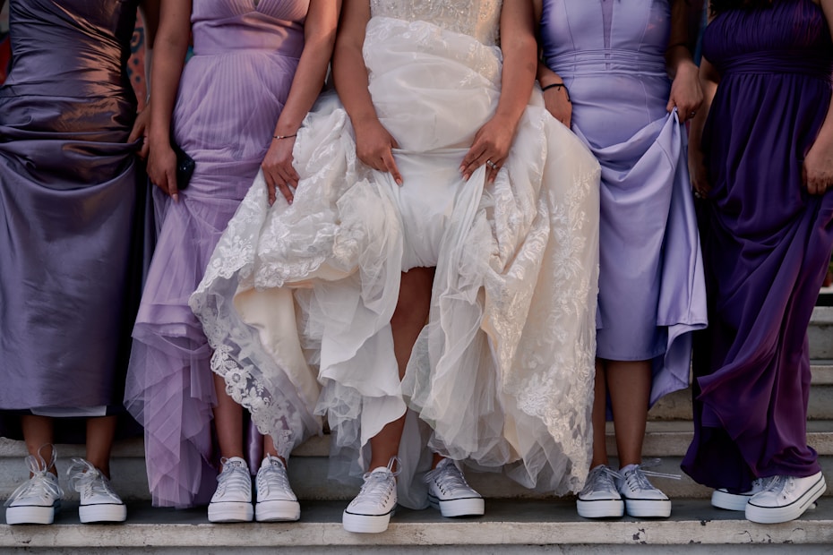 Five women are standing in a line, wearing elegant dresses in shades of purple and lavender with silver sneakers. The central figure is wearing a white lace wedding gown, also paired with sneakers. They are lifting their dresses slightly to display the footwear, standing on stone steps.