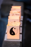 A series of lit paper lanterns featuring a prominent yin-yang symbol on the nearest one are aligned in a row. Each lantern emits a warm glow, and they are placed on small wooden bases on a stone surface.