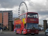 A red double-decker tour bus with colorful designs and the word 'Liverpool' prominently displayed on it is parked on a street. The bus is filled with passengers in the upper deck. In the background, a large Ferris wheel can be seen along with a modern building with the name 'Jury's Inn' and some parked cars.