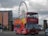A red double-decker tour bus with colorful designs and the word 'Liverpool' prominently displayed on it is parked on a street. The bus is filled with passengers in the upper deck. In the background, a large Ferris wheel can be seen along with a modern building with the name 'Jury's Inn' and some parked cars.