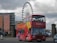 A red double-decker tour bus with colorful designs and the word 'Liverpool' prominently displayed on it is parked on a street. The bus is filled with passengers in the upper deck. In the background, a large Ferris wheel can be seen along with a modern building with the name 'Jury's Inn' and some parked cars.