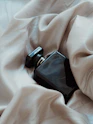 Close-up of a sleek black perfume bottle resting on a softly lit charcoal fabric, accented with faint gold shimmer.