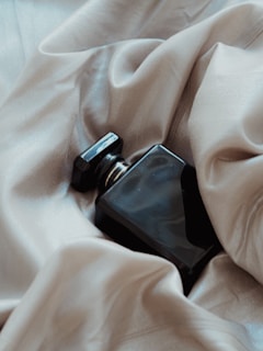 A sleek black perfume bottle rests amid soft, luxurious beige fabric, creating a contrast between the smooth glass and the gentle folds of the cloth.