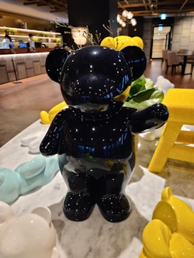 A glossy black bear sculpture is the focal point, surrounded by smaller colorful bear figures in shades of yellow, white, and light blue. They are placed on a marble table in a modern, stylish interior space with wood and metal elements, ambient lighting, and greenery in the background.