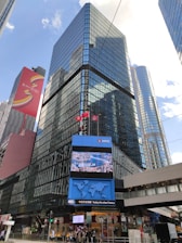 A modern cityscape featuring tall, glass-covered skyscrapers reflecting the blue sky. The buildings are adorned with electronic displays and advertisements. In the foreground, a digital screen shows a map and other information. The street level displays various traffic signs and a group of people near a pedestrian crossing.