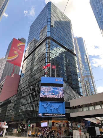 A modern cityscape featuring tall, glass-covered skyscrapers reflecting the blue sky. The buildings are adorned with electronic displays and advertisements. In the foreground, a digital screen shows a map and other information. The street level displays various traffic signs and a group of people near a pedestrian crossing.