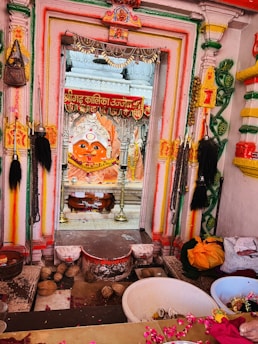 A colorful and intricately decorated temple or shrine entrance adorned with various religious symbols and items. The central focus is a vividly painted idol with a large, striking face. The area is surrounded by garlands, flower petals, and ritualistic objects, creating a vibrant and spiritual atmosphere.