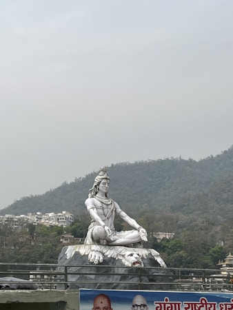 A large statue of a meditative figure sitting cross-legged atop a platform designed like a large tiger skin. The background includes a mountainous landscape with overcast skies and scattered buildings. A partial billboard with text and images is visible at the bottom of the scene.