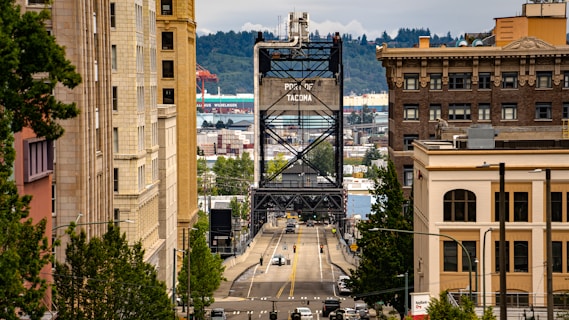 A bustling urban street lined with tall buildings leads to a steel bridge with the words 'Port of Tacoma' visible. Trees and greenery are present along the sidewalks. In the background, there are industrial buildings and shipping containers, indicating a port area. The sky above is overcast.