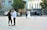 A wedding scene in a city square. A couple dressed in wedding attire stands together with a photographer taking pictures of them. In the foreground, a young man and a young woman are engaged in a conversation. The setting includes ornate buildings and some cars parked along the street.