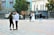 A wedding scene in a city square. A couple dressed in wedding attire stands together with a photographer taking pictures of them. In the foreground, a young man and a young woman are engaged in a conversation. The setting includes ornate buildings and some cars parked along the street.