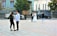 A wedding scene in a city square. A couple dressed in wedding attire stands together with a photographer taking pictures of them. In the foreground, a young man and a young woman are engaged in a conversation. The setting includes ornate buildings and some cars parked along the street.