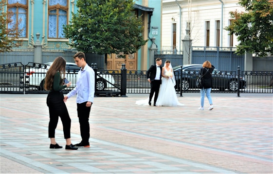 A wedding scene in a city square. A couple dressed in wedding attire stands together with a photographer taking pictures of them. In the foreground, a young man and a young woman are engaged in a conversation. The setting includes ornate buildings and some cars parked along the street.