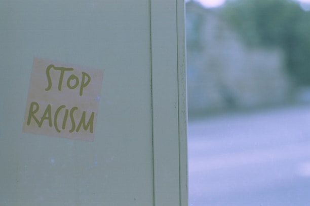 A muted green wall with a paper attached, which reads 'Stop Racism' in green letters. The background is slightly blurred, suggesting an outdoor setting with natural elements like trees.
