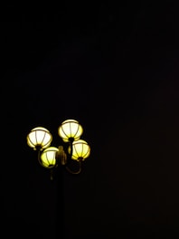 Four spherical street lamps glow with a warm yellow light against a dark, almost black background. The lamps' design is simple yet elegant, encased in a stylish metal frame.