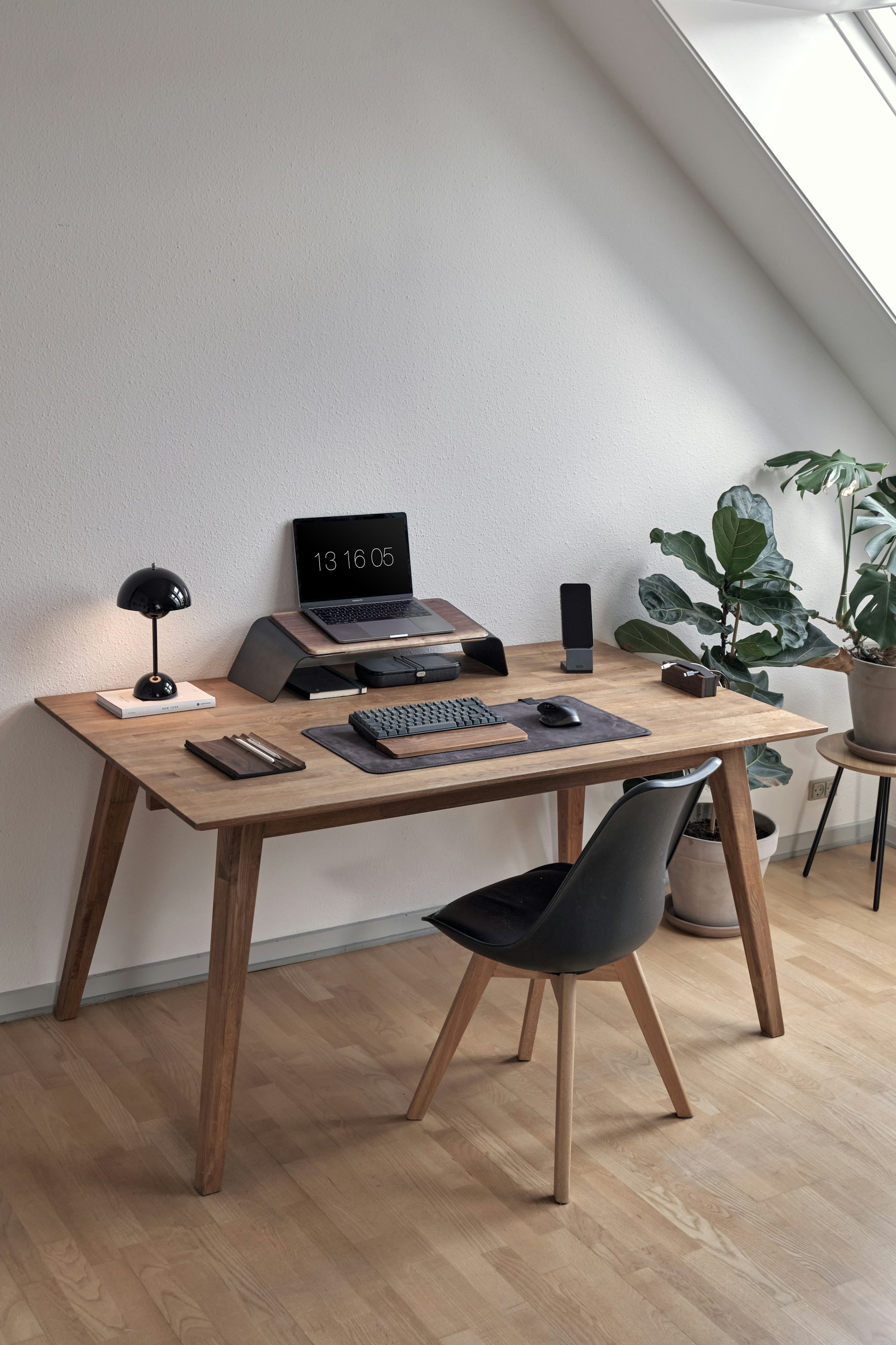 A minimalist home office setup featuring a wooden desk with a modern black chair. On the desk, there is a laptop displaying the time, a keyboard, a mouse, and several desk organizers. A small desk lamp and a potted plant add touches of decor. The space is well-lit by natural light coming through a large window, creating a clean and organized workspace.