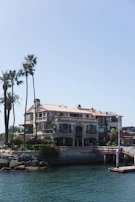 A large, multi-story house with a beige facade is situated by the water. Palm trees are visible beside the house, adding a tropical feel. A wooden dock extends out into the water, and there are rocks lining the shore. The sky is clear and blue.