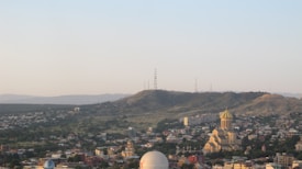A sprawling cityscape features a prominent cathedral with a golden dome, set against a backdrop of rolling hills. The horizon shows a series of hills with communication towers, while the foreground is densely populated with residential buildings and greenery.