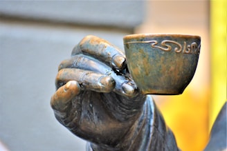 A close-up of a bronze statue hand holding a small cup. The fingers are detailed, and the cup features ornate patterns on its surface with a weathered bronze appearance.
