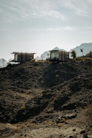 Two modern, rectangular cabins are perched on a rugged, rocky hillside with sparse vegetation. The background features misty mountain silhouettes under a clear, blue sky.