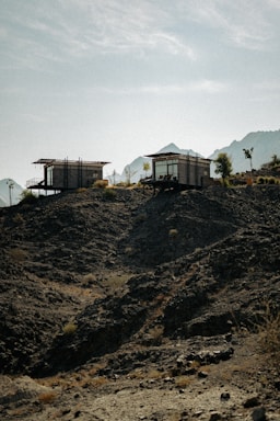 Two modern, rectangular cabins are perched on a rugged, rocky hillside with sparse vegetation. The background features misty mountain silhouettes under a clear, blue sky.