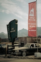 A vintage parking lot with two old cars parked near a weathered wooden building. A large green sign reads 'Hatta Wadi Hub' with the word 'Parking' displayed vertically. A tall red flag with Arabic text and a shopping bag graphic flutters in the wind. In the background, there are mountains and a cloudy sky.