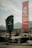 A vintage parking lot with two old cars parked near a weathered wooden building. A large green sign reads 'Hatta Wadi Hub' with the word 'Parking' displayed vertically. A tall red flag with Arabic text and a shopping bag graphic flutters in the wind. In the background, there are mountains and a cloudy sky.