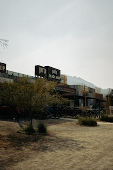 A rustic outdoor setting with a row of small shops or stalls. The signs display 'Hatta Wadi Hub' on top of the storefronts. There are numerous bicycles parked outside, suggesting a recreational or tourist area. The landscape includes dry, sandy terrain and sparse vegetation, with a backdrop of distant mountains under a hazy sky.
