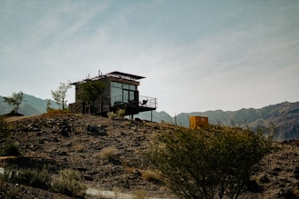 A modern, small cabin with large glass windows is situated on an arid hillside, surrounded by scattered shrubs and sparse vegetation. The structure has a minimalist design, complemented by a metal railing on the balcony. The backdrop features rugged mountains under a clear blue sky, adding to the remote and tranquil ambiance.