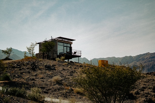 A modern, small cabin with large glass windows is situated on an arid hillside, surrounded by scattered shrubs and sparse vegetation. The structure has a minimalist design, complemented by a metal railing on the balcony. The backdrop features rugged mountains under a clear blue sky, adding to the remote and tranquil ambiance.