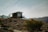 A modern, small cabin with large glass windows is situated on an arid hillside, surrounded by scattered shrubs and sparse vegetation. The structure has a minimalist design, complemented by a metal railing on the balcony. The backdrop features rugged mountains under a clear blue sky, adding to the remote and tranquil ambiance.