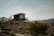 A modern, small cabin with large glass windows is situated on an arid hillside, surrounded by scattered shrubs and sparse vegetation. The structure has a minimalist design, complemented by a metal railing on the balcony. The backdrop features rugged mountains under a clear blue sky, adding to the remote and tranquil ambiance.