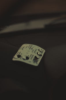 A blister pack of pills with a few tablets removed is resting on a dark surface. The pack has some text written on it, and the lighting is dim, creating a shadowy atmosphere.