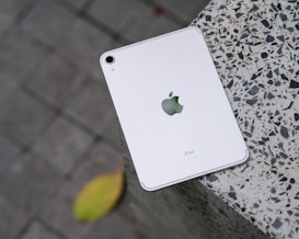 A white iPad with an Apple logo placed on a speckled stone surface. The device is positioned slightly off-center, with a blurred leaf in the background on a gray tiled floor.