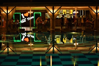 A nighttime scene at a club with an inviting and illuminated bar area featuring a neon sign reading 'I ❤ Sea Time.' Several groups of people are seated around tables, some engaged in conversation, with a pool in the foreground reflecting the lights and activities.
