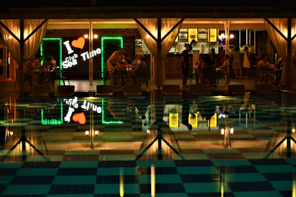 A nighttime scene at a club with an inviting and illuminated bar area featuring a neon sign reading 'I ❤ Sea Time.' Several groups of people are seated around tables, some engaged in conversation, with a pool in the foreground reflecting the lights and activities.