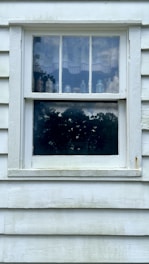 A vintage wooden window with a worn-out, white frame and four-paneled glass. Various glass bottles are visible behind the top glass panes, and a curtain with a scalloped edge partially covers the view. The bottom pane reflects trees and the sky, creating a mirror-like effect.