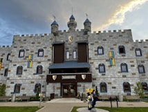 A medieval-style castle with large stone blocks, decorated with colorful banners featuring shields. The castle has multiple small windows with blue and brown accents, and towers on the top with flagpoles. In front of the entrance, there is a sculpture that appears to be a knight dressed in blue and yellow armor. The sky above is partly cloudy with a hint of sunlight breaking through.