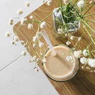A glass of light brown iced coffee or tea with a straw sits on a wooden table. Next to it, a vase holds delicate white flowers with thin green stems, adding a natural touch to the scene.