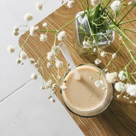A glass of light brown iced coffee or tea with a straw sits on a wooden table. Next to it, a vase holds delicate white flowers with thin green stems, adding a natural touch to the scene.