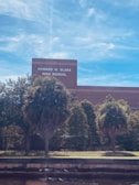 A building with the sign 'Howard W. Blake High School' is visible. The foreground has several palm trees and shrubs, with a fence running parallel to the building. The sky is clear with a few wispy clouds.