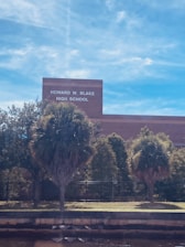 A building with the sign 'Howard W. Blake High School' is visible. The foreground has several palm trees and shrubs, with a fence running parallel to the building. The sky is clear with a few wispy clouds.